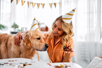 Birthday party. Happy female owner with blond hair presenting golden retriever with small cake and...