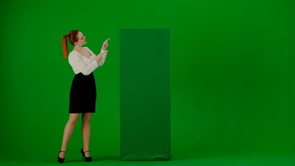 Portrait of attractive office girl on chroma key green screen. Woman in skirt and blouse standing pointing at green advertisement board, smiling.