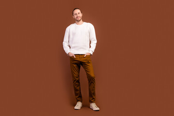 Fototapeta na wymiar Full length photo of good mood cheerful man wear white sweater smiling empty space isolated brown color background