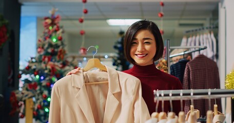 Portrait of happy woman browsing through elegant blazers on racks in Christmas adorn clothing store during winter holiday season. Smiling client checking fashion boutique items