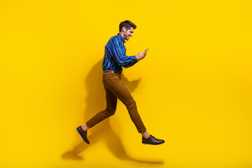 Fototapeta na wymiar Side profile full size photo of busy guy dressed striped shirt brown trousers run look at smartphone isolated on yellow color background