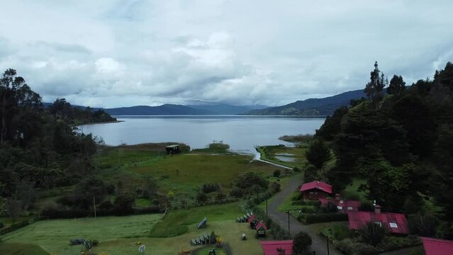 Aerial view of a serene lagoon (La Cocha, Colombia) on a cloudy day, showcasing the roofs of houses surrounding the lagoon and mountains on the horizon