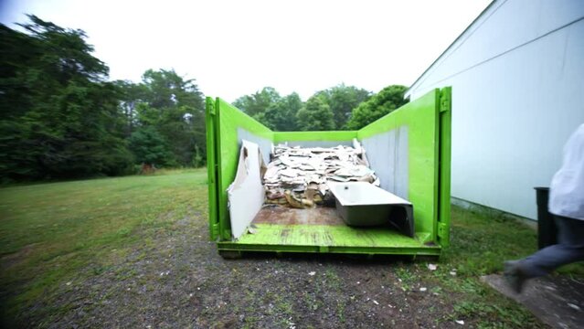 Construction worker wearing respirator doing manual labor carrying pieces of broken wallboard into a dumpster for residential renovation.