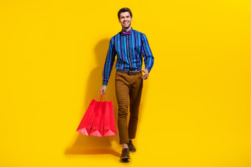 Full size photo of cool cheerful guy dressed striped shirt walking with shopping bags buy new outfit isolated on yellow color background