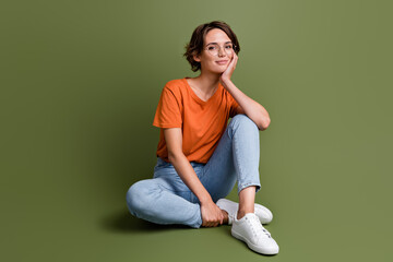 Full length photo of positive cheerful girl wear stylish clothes sit on floor hand on cheekbone...