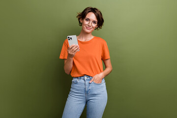 Photo of intelligent cheerful girl with bob hairdo dressed orange t-shirt in glasses hold smartphone isolated on khaki color background