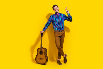 Full size photo of positive guy dressed striped shirt stand with acoustic guitar showing v-sign...