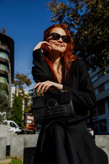 Young and beautiful model poses for the camera. Red-haired model posing for the camera with her crocodile leather, black, luxury bag outside. Fashion photography