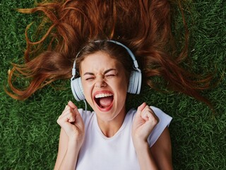 Happy teenage girl listening to music with headphones lying on the green lawn grass in the park, summer vacation