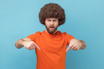 Portrait of man with Afro hairstyle wearing orange T-shirt pointing fingers down commanding to act here and now, demanding immediate result. Indoor studio shot isolated on blue background. - obrazy, fototapety, plakaty
