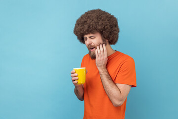 Portrait of man with Afro hairstyle wearing orange T-shirt suffering from terrible teeth pain after drinking hot or cold beverage, dental injury. Indoor studio shot isolated on blue background. - obrazy, fototapety, plakaty