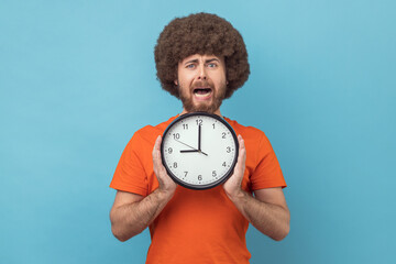 Look at time. Man with Afro hairstyle in orange T-shirt holding big clock and looking displeased...