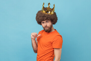 Portrait of serious man with Afro hairstyle wearing orange T-shirt pointing himself, looking at camera with smile, superior privileged status. Indoor studio shot isolated on blue background. - obrazy, fototapety, plakaty