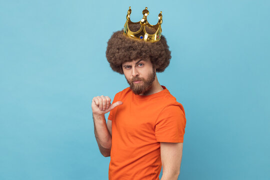 Portrait of serious man with Afro hairstyle wearing orange T-shirt pointing himself, looking at camera with smile, superior privileged status. Indoor studio shot isolated on blue background.