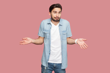Portrait of unaware questioned man standing spreads palms, shrugs shoulders with perplexed expression, being indecisive, has no idea what happened. Indoor studio shot isolated on pink background. - obrazy, fototapety, plakaty