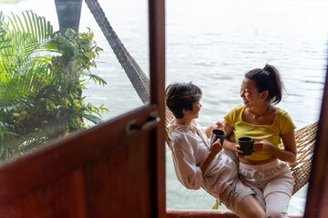 Obraz premium Happy Asian LGBTQ couple relaxing on hammock together at lake house balcony. People enjoy and fun outdoor lifestyle travel nature on summer holiday vacation. LGBT person and pride day concept.