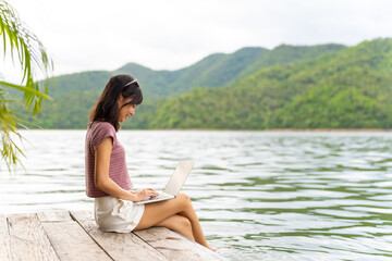 Happy Asian woman sitting on lake house balcony and using laptop computer remote working online business. Attractive girl relax and enjoy outdoor lifestyle travel nature on summer holiday vacation.