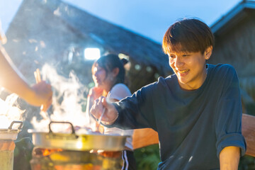 Group of Diversity Asian man and woman friends enjoy and fun celebration meeting dinner party eating barbecue grill and drinking beer together on lake house balcony on summer holiday vacation at night