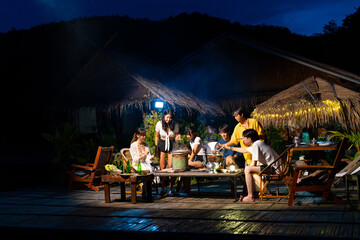 Group of Diversity Asian man and woman friends enjoy and fun celebration meeting dinner party eating barbecue grill and drinking beer together on lake house balcony on summer holiday vacation at night