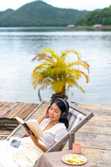 Fototapeta na wymiar Asian woman sitting on lake house balcony reading a book and listening to the music on headphones with mobile app. Attractive girl enjoy outdoor lifestyle travel nature on summer holiday vacation.