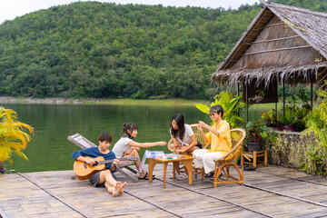 Young Asian man and woman enjoy outdoor lifestyle travel nature forest mountain on summer holiday vacation. Happy generation z people friends playing guitar and doing hobbies at lake house balcony.