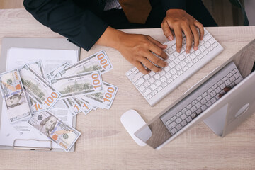 Top view of hands typing on keyboard computer with dollar bills money scattered on the desk