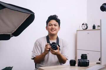 Portrait of young Asian photographer smiling while holding DSLR camera, sitting at photo and video production office.