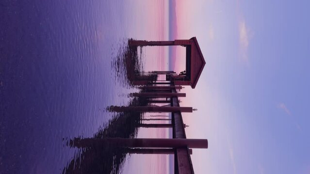 Vertical shot of small pier located on lake shore. Magic sunset landscape. Ripples flowing on water surface. Dusk lights reflecting on lake. Empty wooden jetty on lake surrounded mountains, 4k footage