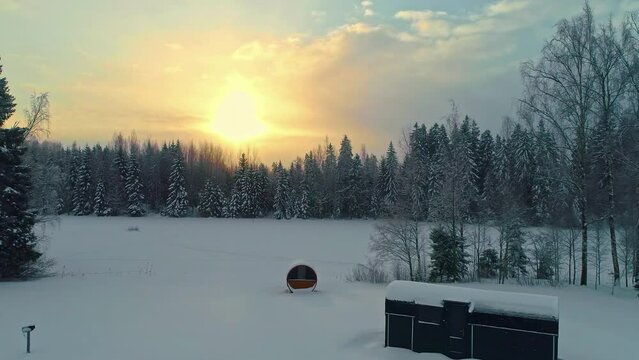 Drone rising backwards over a distant lakeside Sauna and a cottage, wintet sunset