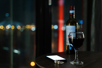 Glass of Red Wine, Smartphone with Glowing Screen, and Wireless Earbuds on Table, Night City Bokeh Lights in Background