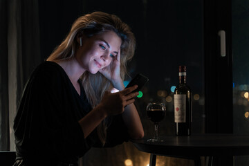 Solitary woman in a night-time interior setting,  engaged with her smartphone while sipping red...