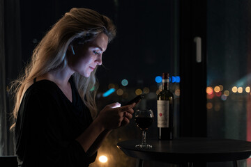 Solitary woman in a night-time interior setting,  engaged with her smartphone while sipping red...