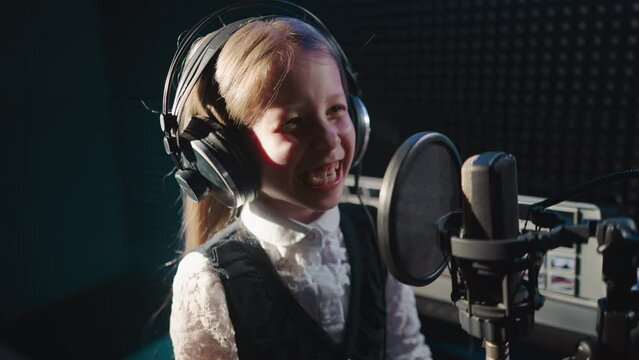 Giggling child in recording studio closeup. Joyful little girl enjoys getting ready for performing song using professional microphone. Soundproof room