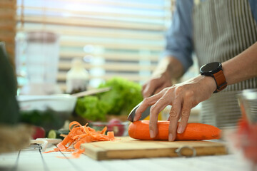 Closeup senior man chopping fresh carrot on board preparing a healthy salad in kitchen.