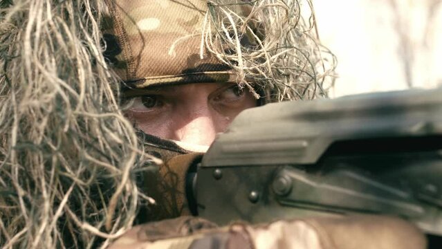 A close-up of a sniper wearing a camouflage suit or grass cloak, waiting and looking through a rifle scope. The sniper aims and shoots. Marksmanship shooting. Cinematic shot