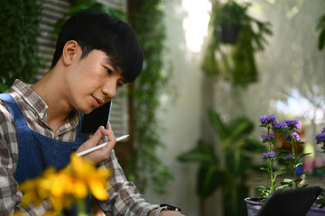 Young male florist checking order on digital tablet during conversation with customer on mobile...
