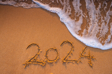 Happy New Year 2024 handwritten number on the beach sand and lapped ocean wave on the shore