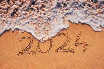 Fototapeta premium New Year 2024 text written on the beach sand and wave at sunrise