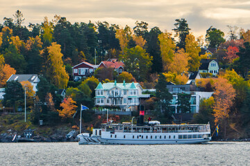 Stockholm, Sweden. Suburbs and residential houses on the islands east of the city in autumn colors. A commuter boat passing by. Colorful trees. - obrazy, fototapety, plakaty