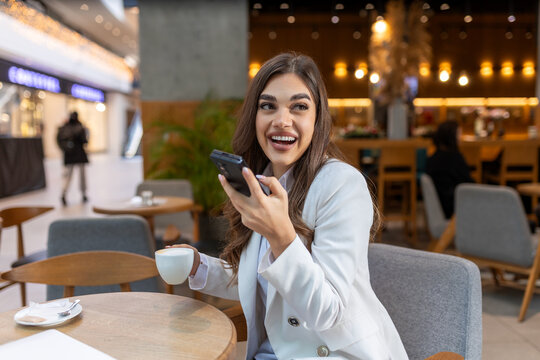 Happy young woman using mobile phone and enjoying coffee at cafe. Young beautiful woman holding coffee cup and looking at smartphone while sitting at cafeteria.