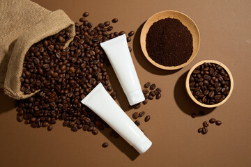 Two cosmetic tubes container cream or mask with coffee extract displayed on brown background with natural ingredient. Coffee beans and grounds on wooden dishes and jute bag
