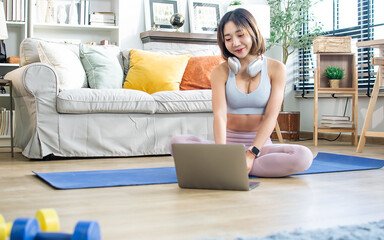 Asian healthy sportive woman wearing sportswear, smiling with happiness, sitting in indoor living room at cozy home, looking at laptop, relax exercising. Recreation, Sport, Lifestyle Concept.