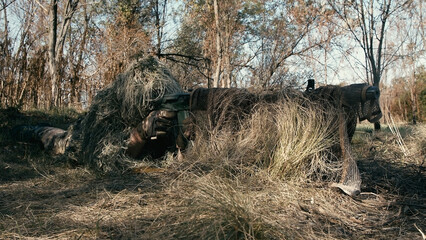 A close-up of a sniper wearing a camouflage suit or grass cloak, waiting and looking through a rifle scope. The sniper aims and shoots. Marksmanship shooting. Cinematic shot