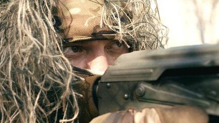 A close-up of a sniper wearing a camouflage suit or grass cloak, waiting and looking through a...