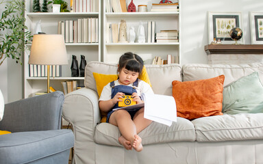 Portrait Asian cute little girl playing alone camera toy with concentration, sitting on sofa in...