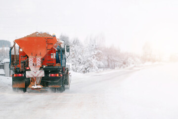 An orange truck with a salt and sand spreader clears the snow and ice from the road. The winter...