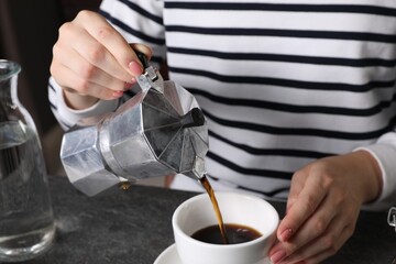 Woman pouring aromatic coffee from moka pot into cup at grey table indoors, closeup