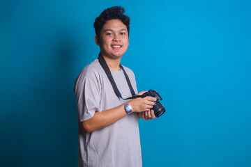 Young photographer standing confidently and smiling while holding camera on blue background