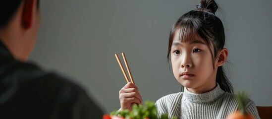 asian girl picky eater turning head and saying no to her dad as he is encouraging her to take a bite of vegetable he holds the food with chopsticks near her. with copy space image - obrazy, fototapety, plakaty