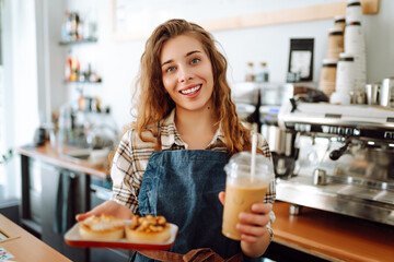 Curly-haired woman barista holding takeaway coffee and dessert. A young woman in an apron behind...